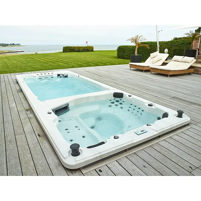 Camargue Hidromasaje para exterior SwimSpa II - 594 x 228 x 137 cm, Gris/Blanco/Marrón, Número de personas máx.: 10 - Vista de una lujosa bañera de hidromasaje con zona de natación integrada, rodeada de una terraza de madera y zonas verdes.