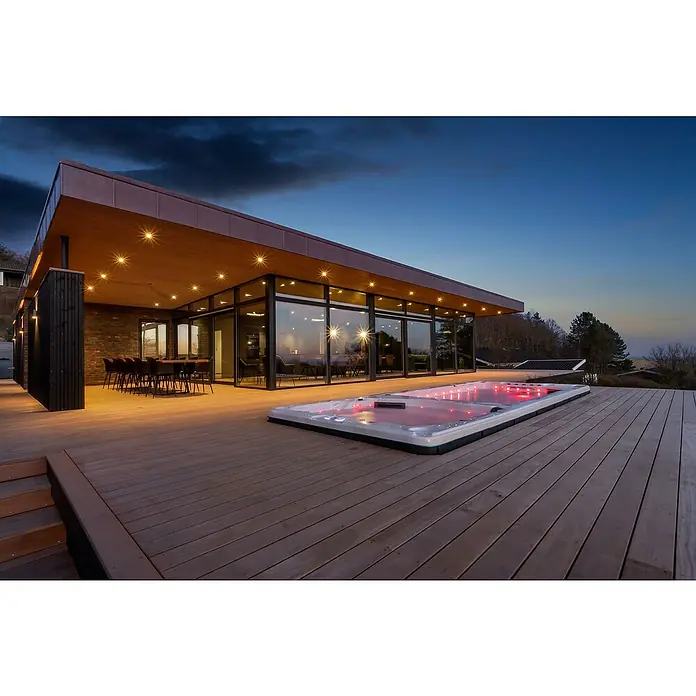Camargue Hidromasaje para exterior SwimSpa II - 594 x 228 x 137 cm, Gris/Blanco/Marrón, Número de personas máx.: 10 - Una lujosa casa moderna con un elegante jacuzzi en la terraza, iluminado con luz roja con un impresionante cielo nocturno.