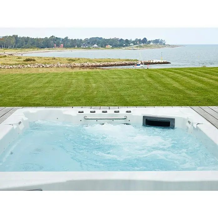 Camargue Hidromasaje para exterior SwimSpa II - 594 x 228 x 137 cm, Gris/Blanco/Marrón, Número de personas máx.: 10 - Una bañera de hidromasaje con agua azul burbujeante en primer plano, con un paisaje verde y costa al fondo.