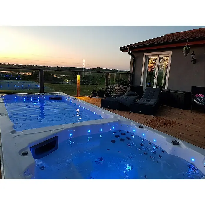 Camargue Hidromasaje para exterior SwimSpa II - 594 x 228 x 137 cm, Gris/Blanco/Marrón, Número de personas máx.: 10 - Lujosa bañera de hidromasaje con iluminación azul en una cubierta de madera al atardecer. Pura relajación en tu propio jardín.