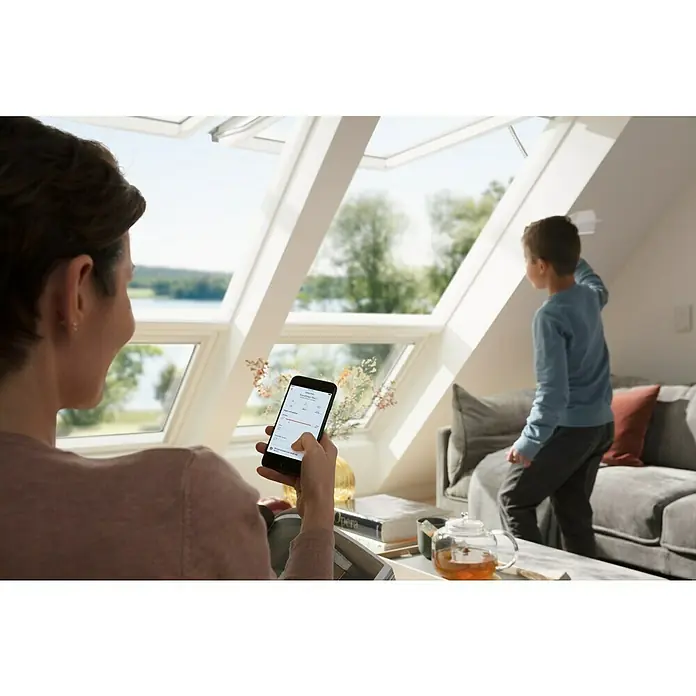 Velux App Control KIG 300 - Passend für: VELUX Solar- & Elektro-Produkte hergestellt seit 2007 - Frau mit Smartphone zur Bedienung von VELUX Dachfenstern, während ein Kind am Fenster steht.