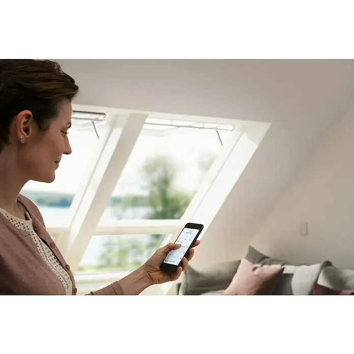 Velux App Control KIG 300 - Passend für: VELUX Solar- & Elektro-Produkte hergestellt seit 2007 - Eine Frau bedient mit einem Smartphone ein VELUX-Produkt in einem Dachzimmer mit Dachfenstern.