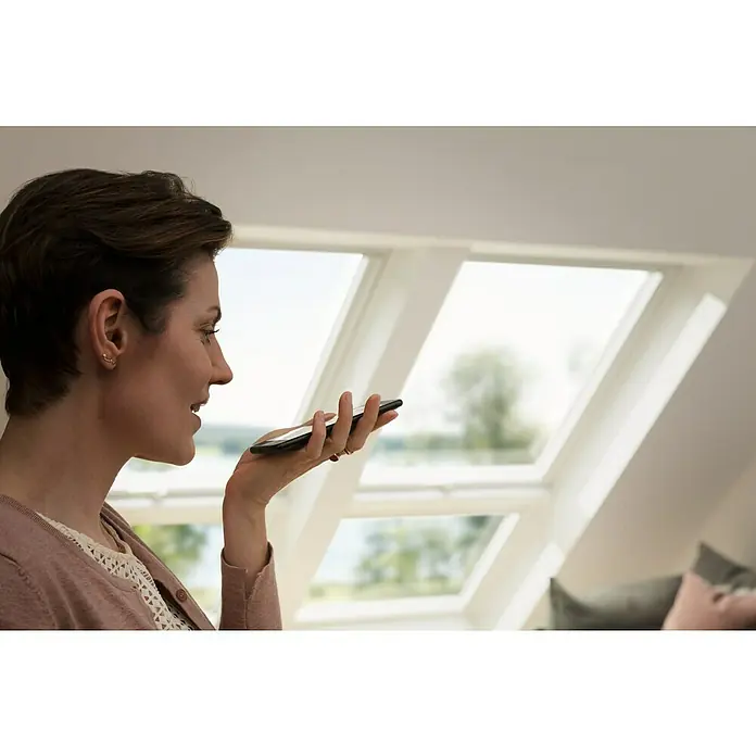 Velux App Control KIG 300 - Passend für: VELUX Solar- & Elektro-Produkte hergestellt seit 2007 - Frau spricht auf ihr Smartphone, ein modernes Gerät für Kommunikation. Durch Dachfenster fällt helles Tageslicht, ideal für klare Sicht.