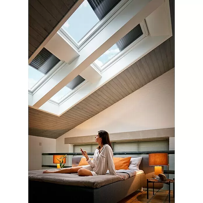 Velux App Control KIG 300 - Passend für: VELUX Solar- & Elektro-Produkte hergestellt seit 2007 - Eine Frau auf einem Bett blickt zu Dachfenstern mit integrierten Jalousien auf und steuert sie mit einem Smartphone.