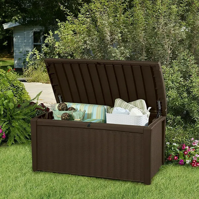 Keter Baúl para jardín Borneo - Plástico, 70 x 130 x 63 cm, 400 l, Marrón - Una caja de almacenamiento para exteriores marrón con tapa abierta en el jardín. La caja está llena de cojines y una tina de almacenamiento blanca.