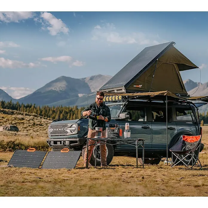 Jackery Panel solar Saga - 80 W, 90,6 x 52,8 cm - Un hombre sirve café junto al generador solar Jackery frente a un Bronco con tienda de techo y paneles solares.