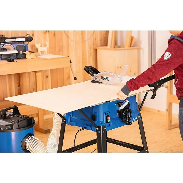 Scheppach Tischkreissäge HS100S - 2.000 W, Durchmesser Sägeblatt: 250 mm, Schnitttiefe bei 90 °: 85 mm - Eine blaue Scheppach Tischkreissäge mit einem Werkstück aus Holz, das von einer Person bearbeitet wird, steht in einer Werkstatt.