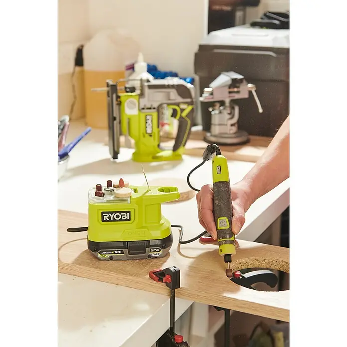 Ryobi ONE+ Akku-Multifunktionswerkzeug RRT18-0 - 18 V, Ohne Akku, Leerlaufdrehzahl: 4.400 U/min - 23.000 U/min - Nahaufnahme eines Ryobi-Rotationswerkzeugs, das zum Schleifen eines Lochs in einem Holzstück verwendet wird, mit Zubehör und Werkzeugen im Hintergrund.