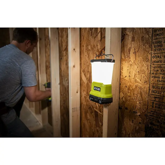 Ryobi ONE+ LED-Leuchte RLL18-0 - 850 lm, Ohne Akku - Akku-betriebene Ryobi Laterne leuchtet eine Baustelle aus, während ein Mann im Hintergrund arbeitet, wodurch die Szene gut beleuchtet ist.