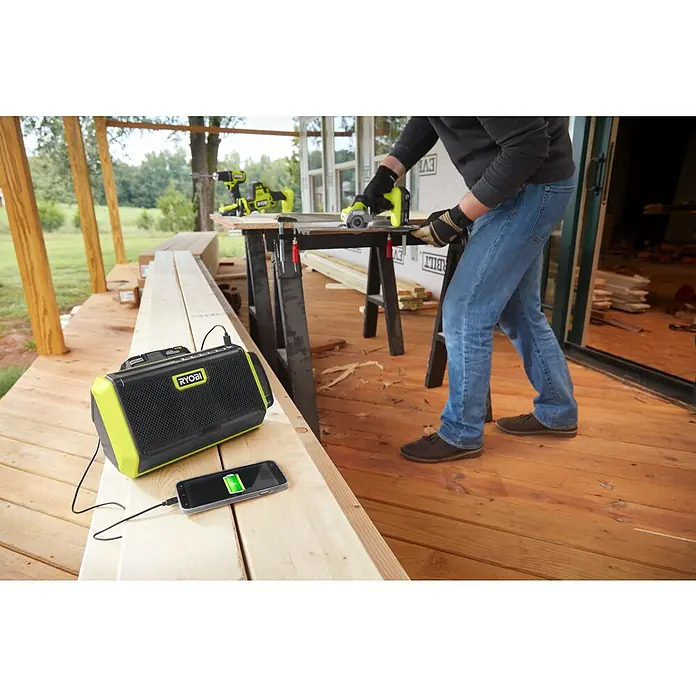 Ryobi ONE+ Altavoz Bluetooth RBT18-0 - 18 V, Sin batería, Potencia de salida altavoz: 15 W - Radio Ryobi con teléfono cargando mientras un hombre trabaja con herramientas al fondo.