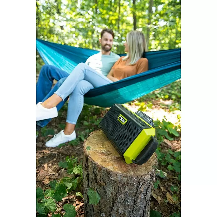 Ryobi ONE+ Altavoz Bluetooth RBT18-0 - 18 V, Sin batería, Potencia de salida altavoz: 15 W - Altavoz Ryobi negro y verde neón sobre un tocón de árbol, con dos personas relajándose en una hamaca en el fondo.