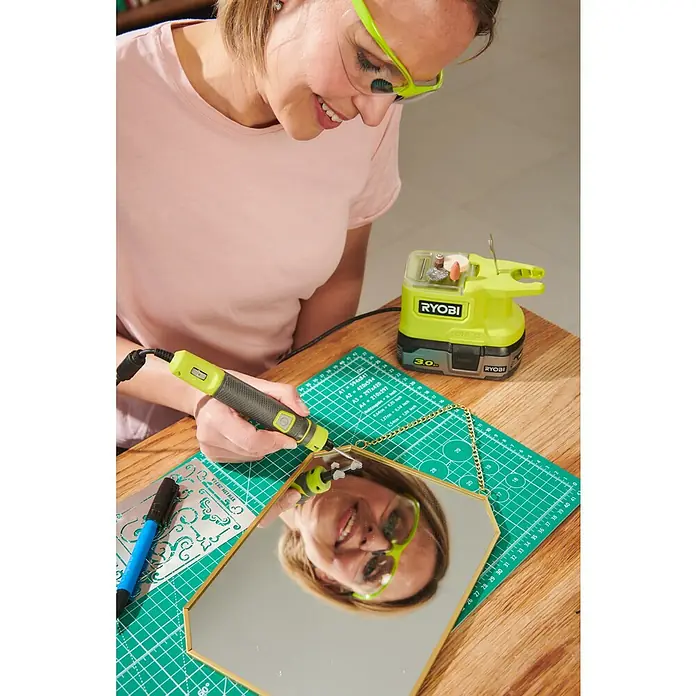 Ryobi ONE+ Akku-Multifunktionswerkzeug RRT18-0 - 18 V, Ohne Akku, Leerlaufdrehzahl: 4.400 U/min - 23.000 U/min - Frau graviert mit einem Ryobi-Rotationswerkzeug ein Spiegelbild auf einem Tisch mit Zubehör.