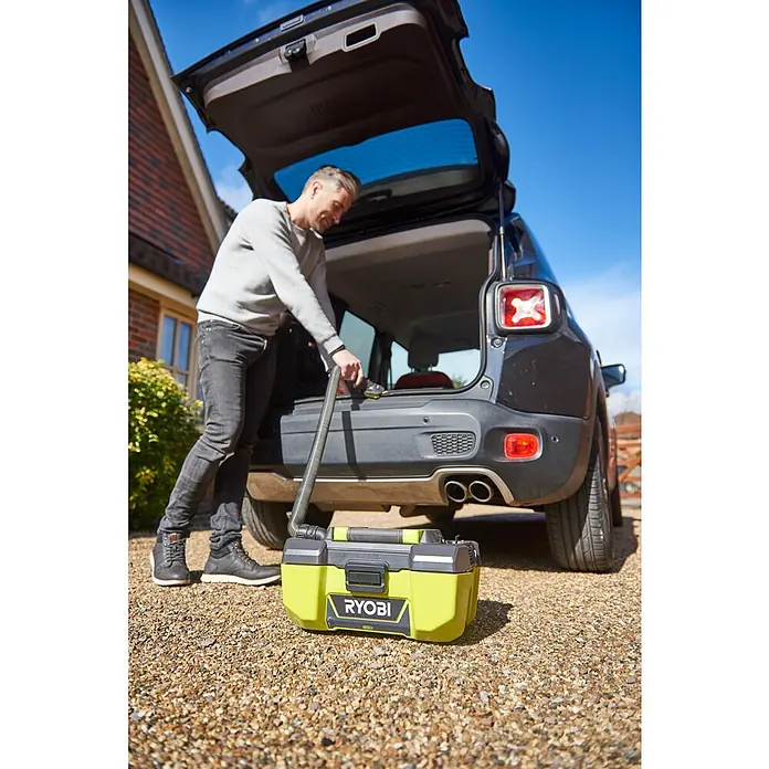 Ryobi Zubehör-Set RAKVA04 - 6 Stk. - Ein Mann verstaut einen gelb-schwarzen RYOBI Akku-Nass-Trockensauger im Kofferraum eines schwarzen Autos vor einem Haus.