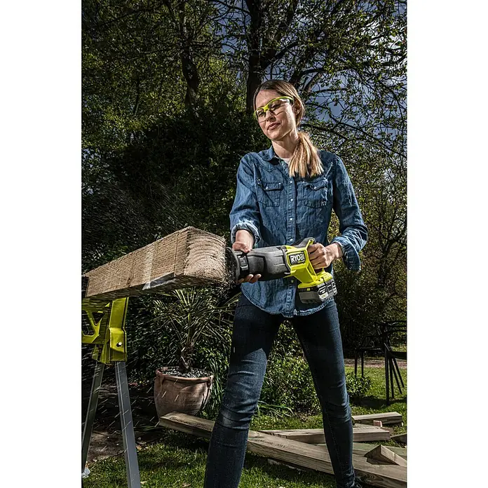 Ryobi ONE+HP Akku-Säbelsäge RRS18X-0 - 18 V, Hublänge: 32 mm, Ohne Akku - Eine Frau sägt im Freien ein Stück Holz mit einer RYOBI Säbelsäge, wobei sie eine Schutzbrille trägt.