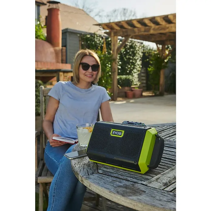 Ryobi ONE+ Altavoz Bluetooth RBT18-0 - 18 V, Sin batería, Potencia de salida altavoz: 15 W - Mujer sentada con una radio RYOBI sobre una mesa de madera al aire libre. La radio es negra con detalles verdes y tiene un diseño robusto.