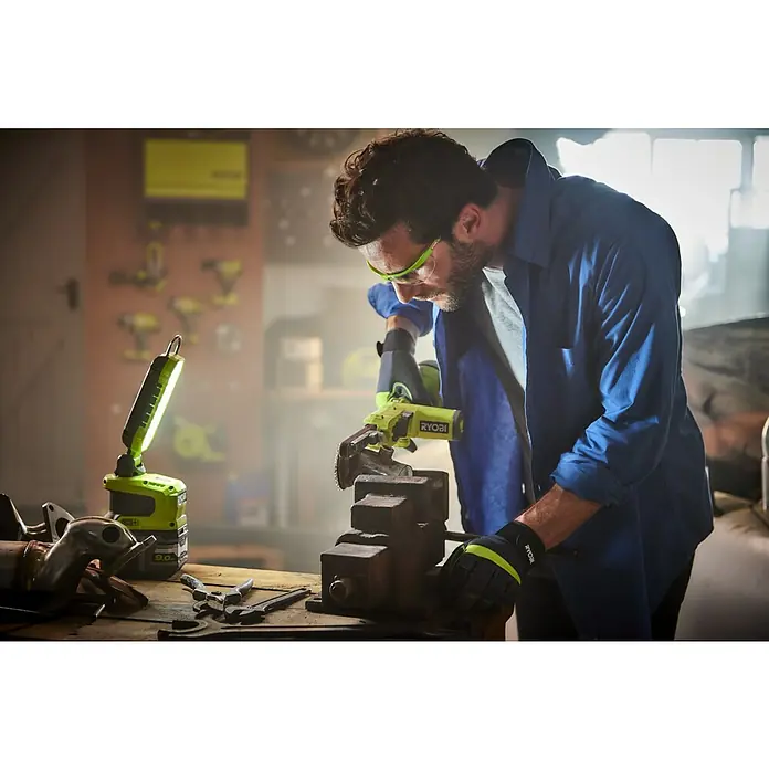 Ryobi ONE+ LED-Arbeitsleuchte R18ALP-0 - 18 V, Li-Ionen, Ohne Akku, 900 lm - Nahaufnahme eines Mannes, der mit einem Ryobi-Werkzeug an einem Schraubstock arbeitet, mit einem Ryobi-Arbeitslicht daneben.