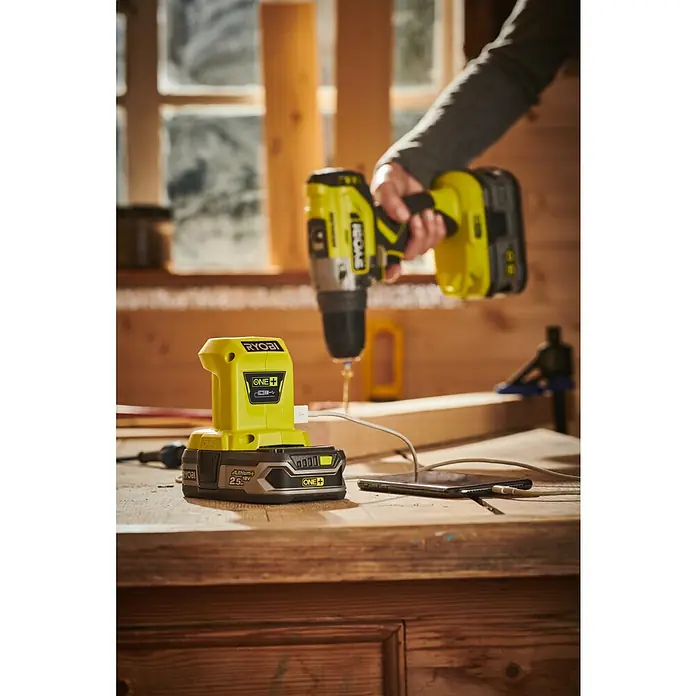 Ryobi ONE+ USB-Adapter - 18 V, Ohne Akku - Nahaufnahme eines Ryobi ONE+ Akkus mit USB-Anschluss, der ein Smartphone auflädt, während ein Ryobi-Bohrer bereit steht.