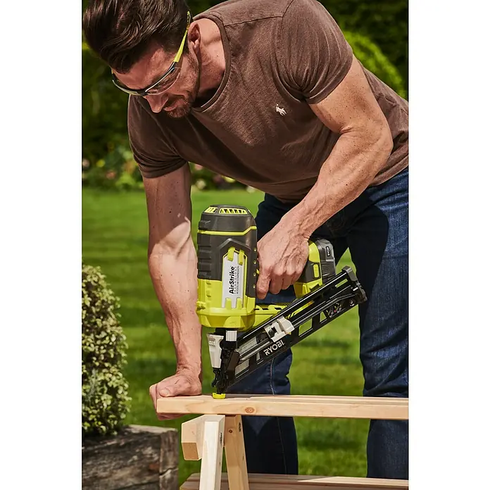 Ryobi ONE+ Akku-Nagler R15GN18-0 - 18 V, Ohne Akku - Ein Mann benutzt einen gelben und schwarzen Ryobi AirStrike Nagler, um ein Holzstück zu befestigen. Sicherheit geht vor, er trägt eine Schutzbrille.