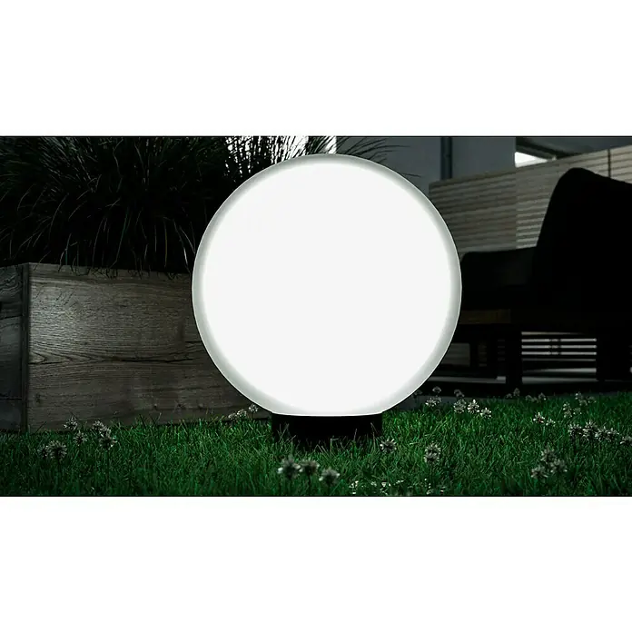 Starlux Kugel-Außenleuchte Kira - 50 cm, Weiß - Außenaufnahme einer leuchtenden Kugel-Gartenleuchte auf einem Rasen, umgeben von Pflanzen und einem Holzpflanzgefäß.
