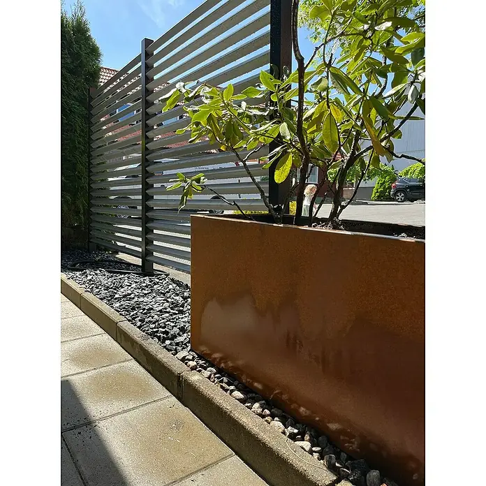 Recal Pflanzkübel eckig - Außenmaß (L x B x H): 120 x 40 x 50 cm, Metall, Braun - Ein modernes Gartendesign mit einem rostfarbenen Pflanzgefäß und einem stilvollen, horizontalen Lattenzaun, umgeben von Zierkies.