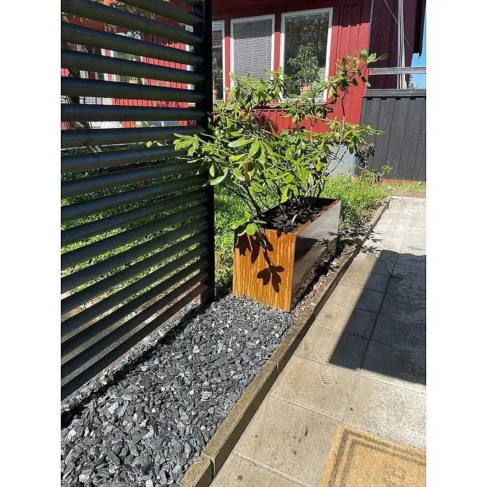 Recal Pflanzkübel eckig - Außenmaß (L x B x H): 120 x 40 x 50 cm, Metall, Braun - Eine moderne Gartenszene mit einem schwarzen Lamellenzaun, einem rostfarbenen Pflanzgefäß und dunklen Ziersteinen.