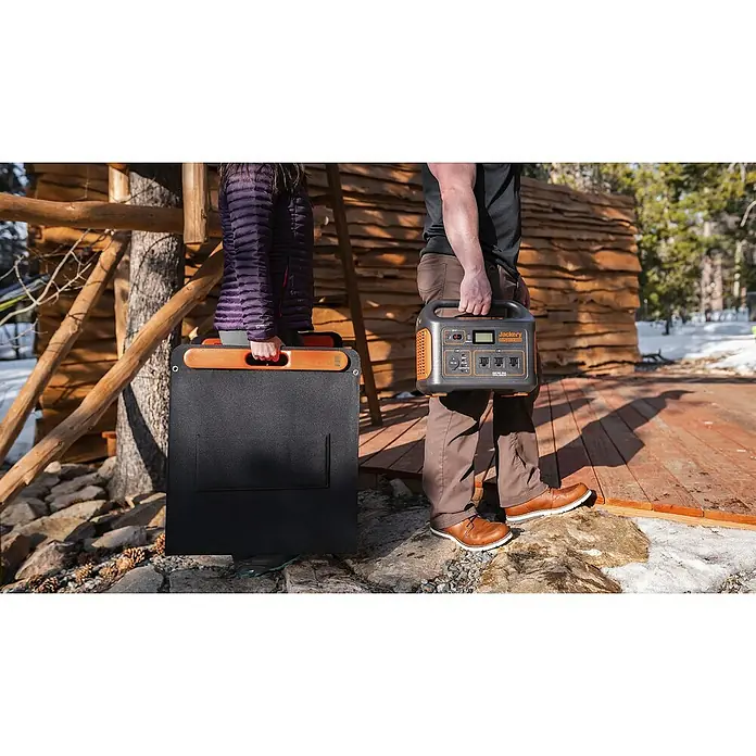 Jackery Colector luz solar Saga - 100 W, 122 x 53,5 cm - Dos personas llevando un panel solar Jackery y una estación de energía Jackery frente a una cabaña de madera. Equipo Jackery ideal para actividades.