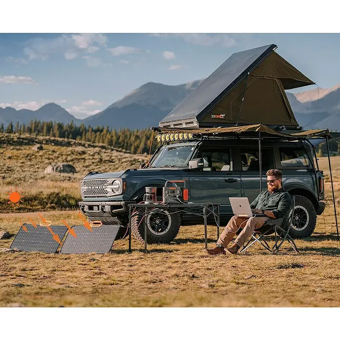 Jackery Panel solar Saga - 80 W, 90,6 x 52,8 cm - Escena de camping con paneles solares y estación de energía Jackery. Un hombre trabaja en un portátil frente a un SUV con tienda de techo.