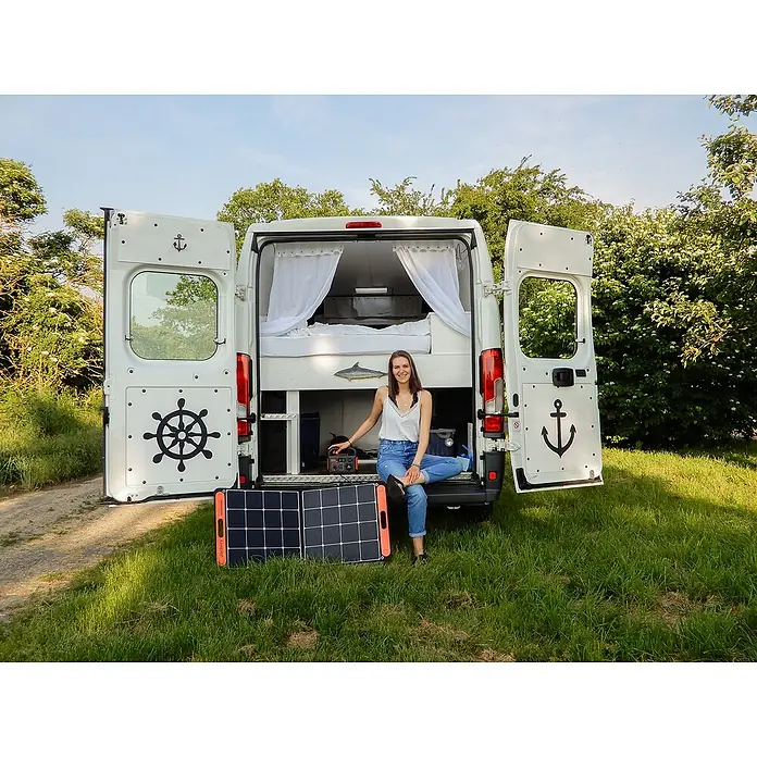 Jackery Estación de energía portátil Explorer 240 Pro EU - 240 Wh, Negro, 16,8 Ah - Mujer sentada frente a una autocaravana con las puertas abiertas y paneles solares Jackery en primer plano.