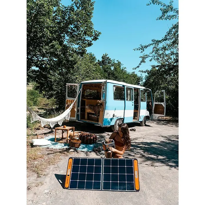 Jackery Estación de energía portátil Explorer 240 Pro EU - 240 Wh, Negro, 16,8 Ah - Una mujer sentada frente a un generador solar Jackery, con una furgoneta azul al fondo. Ideal para viajes de camping y aventuras al aire libre.
