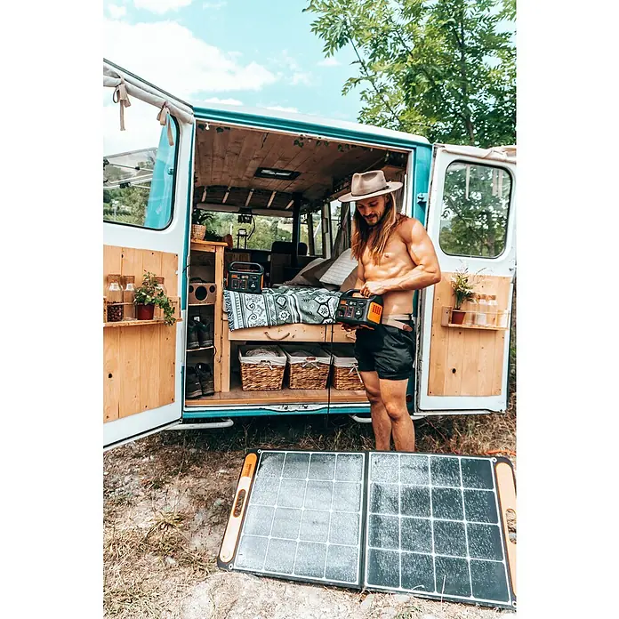 Jackery Estación de energía portátil Explorer 240 Pro EU - 240 Wh, Negro, 16,8 Ah - Un hombre con sombrero y torso desnudo opera una estación de energía Jackery frente a una caravana con panel solar.