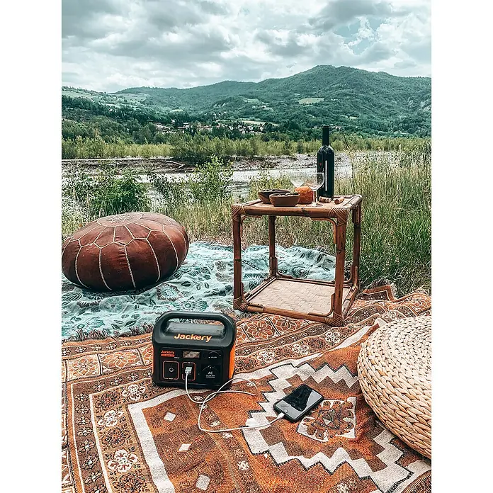 Jackery Estación de energía portátil Explorer 240 Pro EU - 240 Wh, Negro, 16,8 Ah - Una central eléctrica Jackery está cargando un teléfono inteligente sobre una manta al aire libre, perfecto para aventuras al aire libre y disfrutar de la naturaleza con energía.