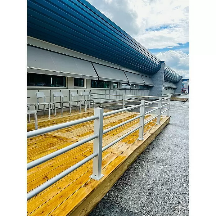 Recal Zaunpfosten Hindsen - Höhe: 99,5 cm, Zwischenpfosten, Natur eloxiert, Aluminium - Eine nasse Holzterrasse mit weißem Metallgeländer, grauen Stühlen und einem modernen Gebäude mit blauen und grauen Fassadenelementen.