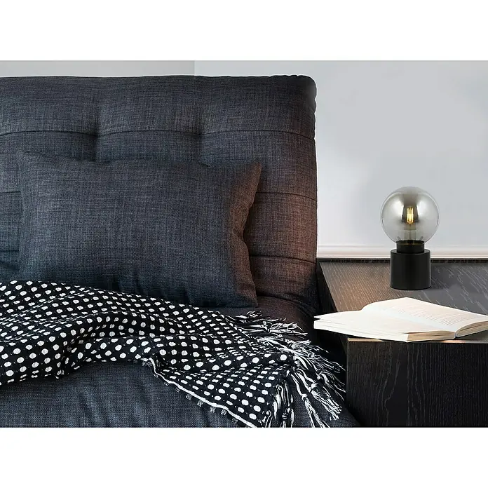 Globo Tischleuchte rund MARKA - 25 W, Ø x H: 15 x 24,5 cm, Schwarz-matt, E14 - Nahaufnahme eines Sofas mit Kissen, einer Decke und einer Lampe auf einem Nachttisch mit Buch, das eine gemütliche Atmosphäre erzeugt.