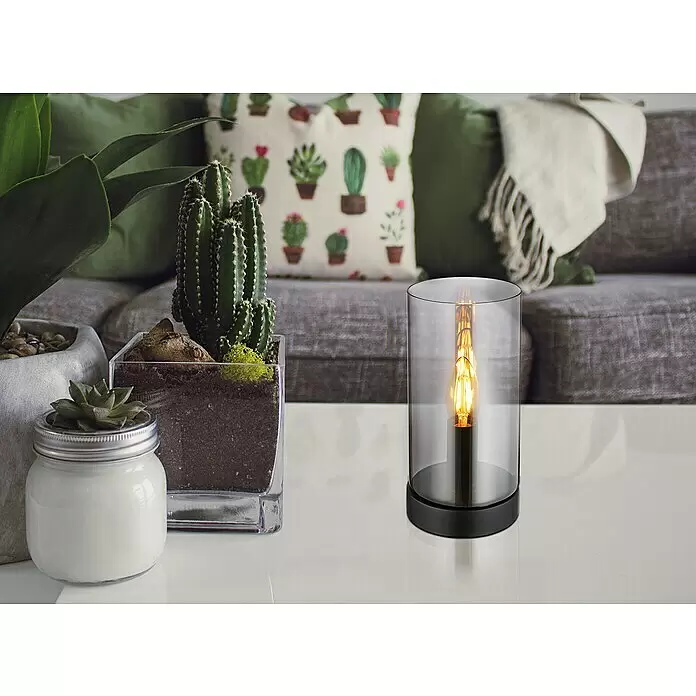 Globo Lámpara de sobremesa redonda Olly - 25 W, Ø x Al: 11 x 22,5 cm, Negro, E14 - Lámpara de mesa cilíndrica con base negra y cristal transparente, con una bombilla de filamento encendida, sobre una mesa blanca en una sala de estar.