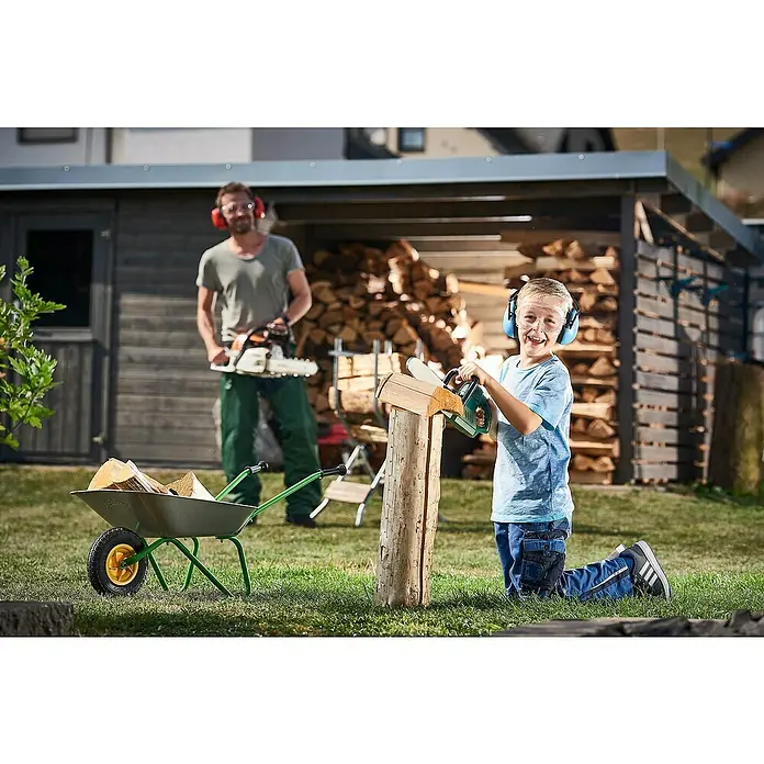 Wolfcraft Schutzbrille Kids - Geeignet für: Kinder - Ein junger Junge sägt mit einer Spielzeugkettensäge Holz, während ein Mann mit einer echten Kettensäge im Hintergrund steht. Beide tragen Ohrenschützer.