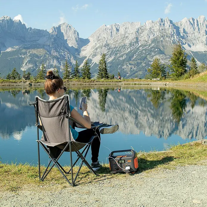 Jackery Estación de energía portátil Explorer 500 Pro EU - 500 Wh, Negro, 24 Ah - Mujer en silla de camping cargando su móvil con una estación de energía Jackery con montañas de fondo.