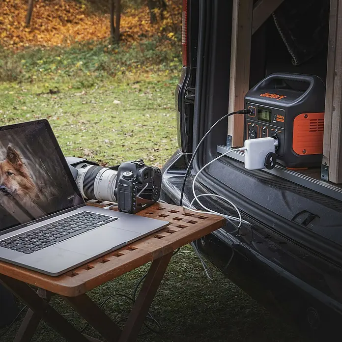 Jackery Estación de energía portátil Explorer 500 Pro EU - 500 Wh, Negro, 24 Ah - Imagen de una estación de energía Jackery conectada a una mesa con un portátil y una cámara, ideal para alimentar dispositivos sobre la marcha.