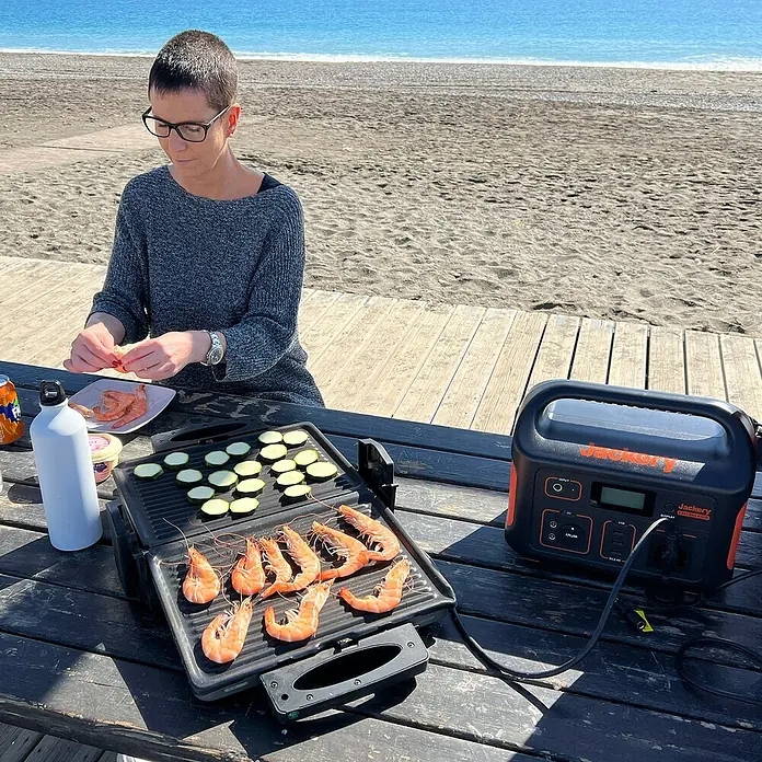 Jackery Estación de energía portátil Explorer 500 Pro EU - 500 Wh, Negro, 24 Ah - Mujer asando gambas y calabacín con una estación de energía Jackery en una playa, ideal para acampar.