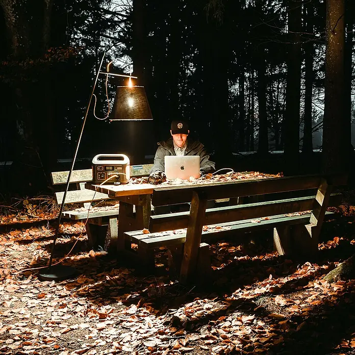 Jackery Estación de energía portátil Explorer 500 Pro EU - 500 Wh, Negro, 24 Ah - Un hombre trabajando en un portátil con una estación de energía y una lámpara en una mesa en el bosque. Alimentación móvil.