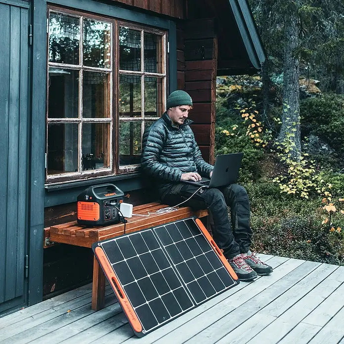 Jackery Estación de energía portátil Explorer 500 Pro EU - 500 Wh, Negro, 24 Ah - Un hombre se sienta con una computadora portátil en un banco junto a un generador solar Jackery y un panel solar frente a una cabaña.