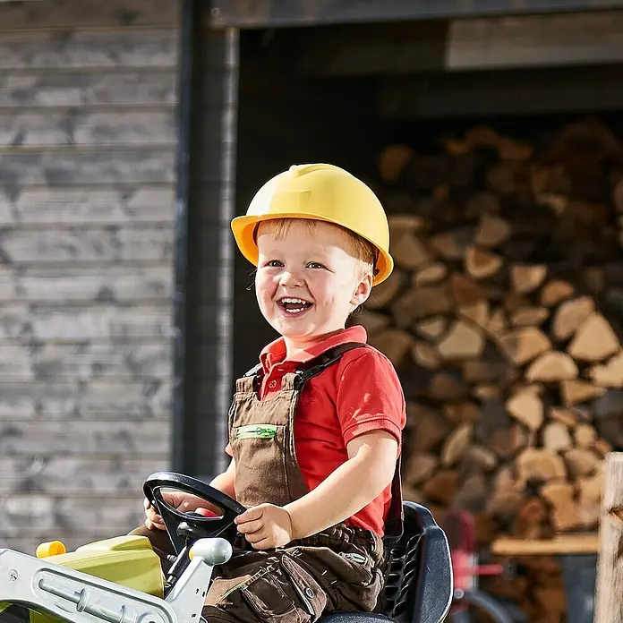 Casco de protección Kids - Amarillo - Un niño sonriente con un casco está sentado en un tractor de juguete delante de madera apilada, irradiando alegría y felicidad.