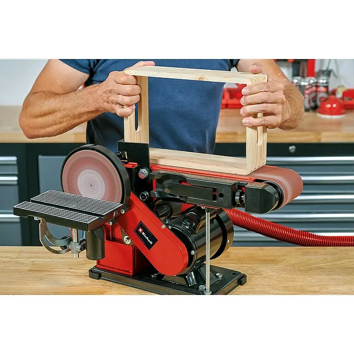 Einhell Lijadora de banda eléctrica de sobremesa TC-US 380 - 380 W, Número de revoluciones de marcha en vacío: 1.500 r.p.m. - Un hombre usa una lijadora de banda Einhell para alisar un marco de madera. La lijadora es roja y negra, y el marco de madera es de color claro.
