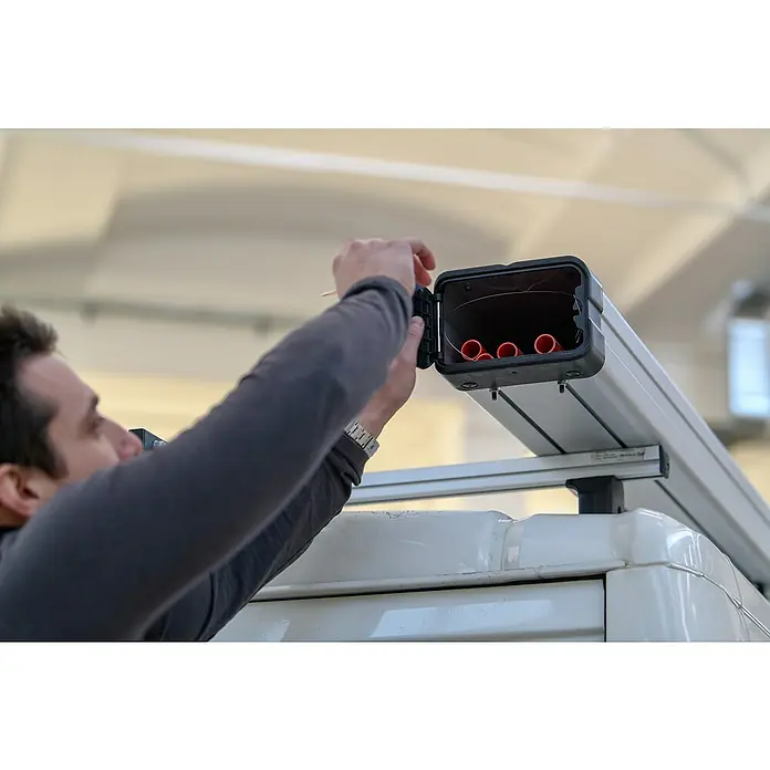Menabo Transportbox Professional für lange Gegenstände - L x B x H: 320 x 15,5 x 21,5 cm, Aluminium - Mann öffnet einen Behälter mit roten Rohren, der an einem weißen Fahrzeugdach befestigt ist. Das Produkt ist ein vielseitiger Dachträger für Fahrzeuge.