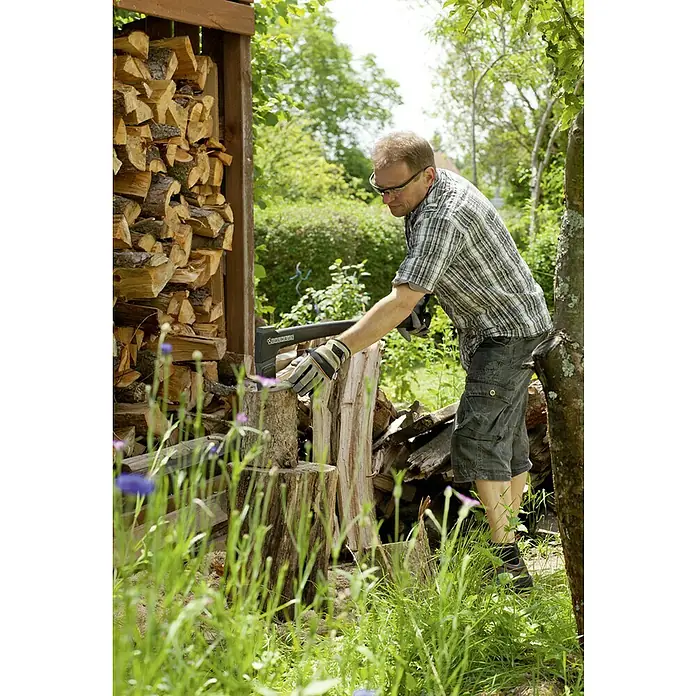 Gardol Profilinie Universalbeil - Länge: 37 cm - Ein Mann spaltet Holz mit einer Axt, Holzscheite liegen bereit. Ein Holzstapel ist im Hintergrund zu sehen, umgeben von Grün.