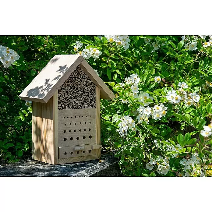 Dobar Insektenhotel Maja - 22,5 x 14 x 29,5 cm, Holz, Natur - Holz-Insektenhotel mit Bambusröhren und Bohrlöchern, im Freien zwischen grünem Laub und weißen Blüten platziert.