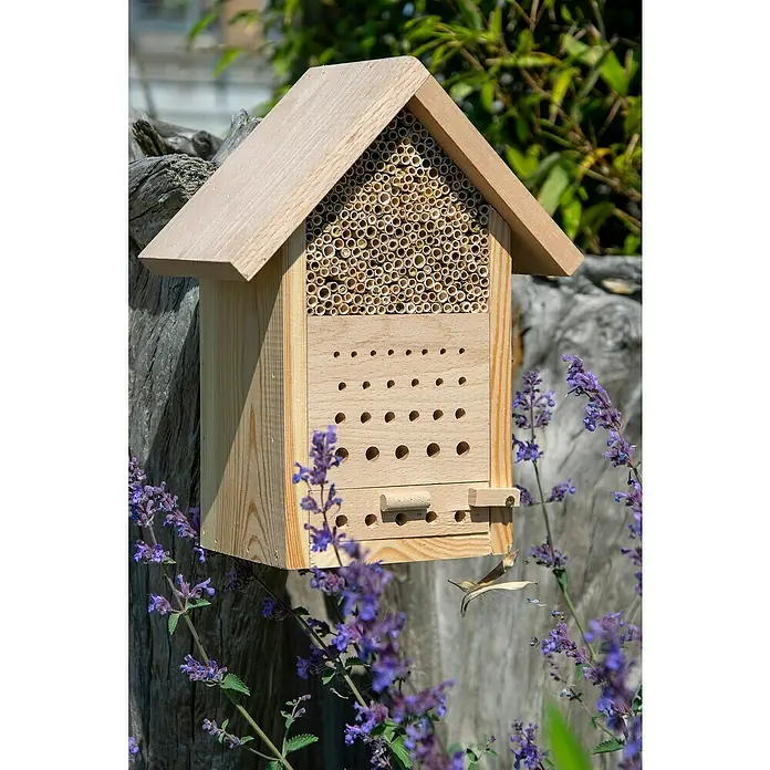 Dobar Insektenhotel Maja - 22,5 x 14 x 29,5 cm, Holz, Natur - Ein hölzernes Insektenhotel hängt an einem Baumstumpf, umgeben von lila Blumen. Das Hotel bietet Nistplätze für verschiedene Insektenarten.
