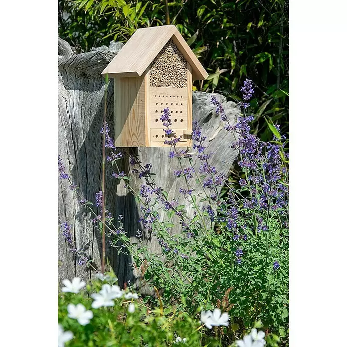 Dobar Bienenhaus Maja - L x B x H: 22,5 x 14 x 29,5 cm, Kiefer - Insektenhotel aus Holz mit Bambusröhrchen, auf einem Baumstumpf inmitten violetter Blumen im Garten.