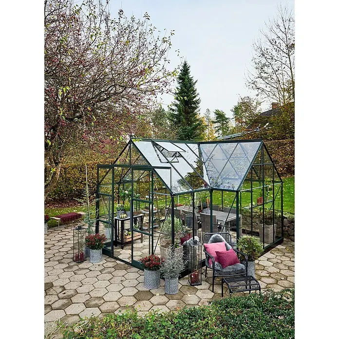 Halls Gewächshaus Garden Room - Außenmaß inkl. Dachüberstand (B x T): 390 x 389 cm, Material Seitenwand: Glas, 3 mm, Grün - Das Bild zeigt ein Gewächshaus mit Glaswänden in einem Garten mit Stühlen und Pflanzen. Das Gewächshaus ist gut beleuchtet.