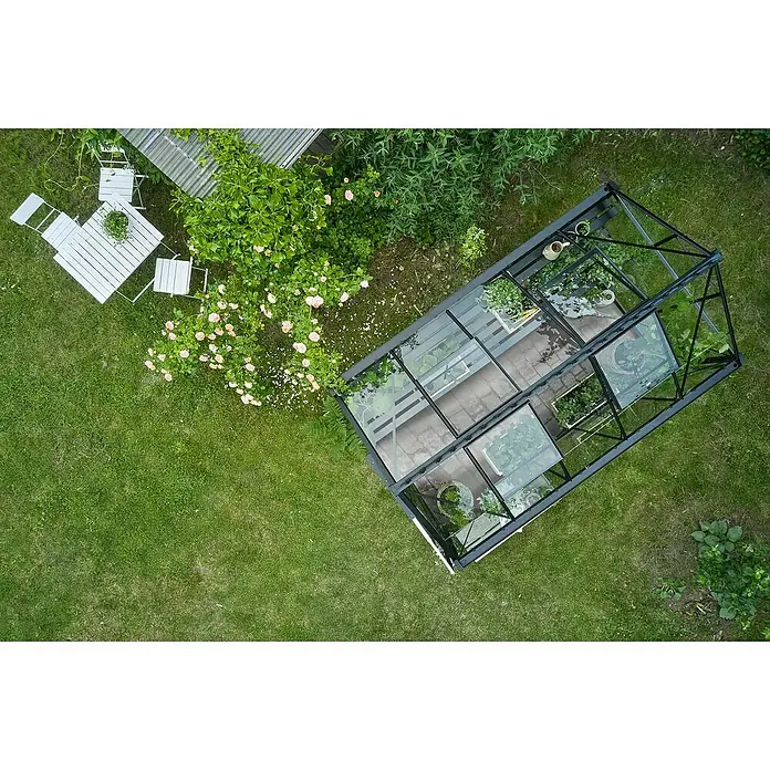 Halls Gewächshaus Burford 610 - Außenmaß inkl. Dachüberstand (B x T): 194 x 318 cm, Material Seitenwand: Glas, 3 mm, Schwarz - Luftaufnahme eines Gewächshauses in einem Garten mit Rasen, Blumen und einer kleinen Sitzecke. Das Gewächshaus beherbergt verschiedene Pflanzen.