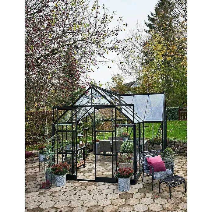 Halls Gewächshaus Garden Room - Außenmaß inkl. Dachüberstand (B x T): 262 x 381 cm, Material Seitenwand: Glas, 3 mm, Schwarz - Das Bild zeigt ein Gewächshaus mit schwarzem Rahmen, das in einem Garten auf einer Terrasse steht und mit Möbeln ausgestattet ist.